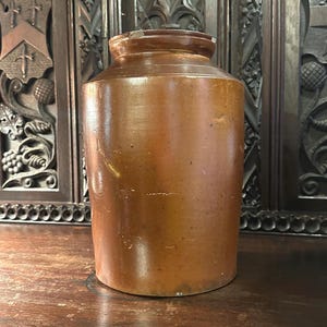 Pode incluir: Um jarro de grés castanho com um corpo cilíndrico e uma borda ligeiramente alargada. O jarro antigo tem uma aparência rústica, com imperfeições visíveis e variações no esmalte. O jarro é exibido em uma superfície de madeira, com um fundo de madeira escura.