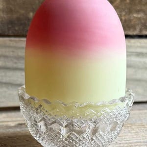 Puede incluir: Una vela decorativa en un portavelas de cristal transparente con diseño de diamantes. La vela tiene un degradado de colores, que va del rojo al rosa y al amarillo. El portavelas de cristal tiene un borde festoneado.