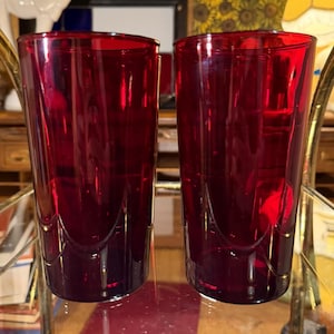 Op de afbeelding: Twee robijnrode drinkglazen. De glazen zijn hoog en cilindrisch met een lichte curve nabij de basis. De glazen zijn gemaakt van een transparant materiaal, waardoor licht erdoorheen kan. De glazen zijn leeg en staan op een reflecterend oppervlak.