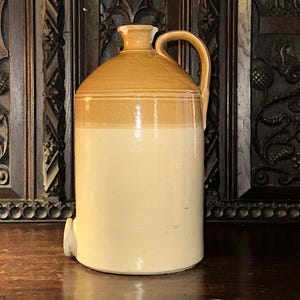May include: A two-tone stoneware jug in cream and tan. The jug has a handle, a spout, and a rounded top. It is likely designed for storing liquids and has a classic, rustic appearance. The jug is displayed on a wooden surface.
