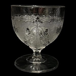 May include: Clear glass goblet with a textured, leaf-patterned bowl and a round base. The stem is short and faceted. The glass has a vintage aesthetic, suitable for serving beverages.