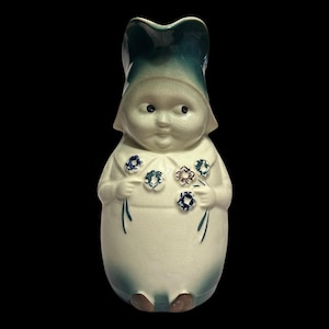 Puede incluir: Figura de cerámica de una figura infantil con un sombrero grande y flores. La figura es principalmente blanca con detalles en azul y marrón. El sombrero es de un tono azul más oscuro. La figura tiene un diseño caprichoso y es probablemente un artículo decorativo.