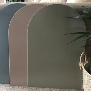 May include: Three arched panels in shades of blue-grey, beige, and olive green. The panels are arranged side by side, with a curved top and a flat bottom. The panels are leaning against a wall, with a plant and a woven basket in the background.