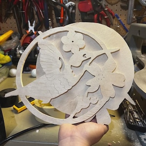 May include: A wooden hummingbird and flower cut-out design. The hummingbird is in flight, with its wings spread. The design is set within a circular frame. The wood is unfinished and light in color.