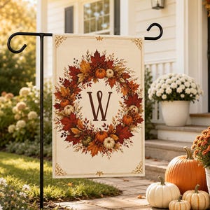 Könnte beinhalten: Eine rechteckige Gartenflagge mit cremefarbenem Hintergrund und einem herbstlichen Kranz aus roten, orangen und gelben Ahornblättern, Beeren und kleinen Kürbissen. In der Mitte ist der Buchstabe W in dunkelbrauner Serifenschrift gedruckt.
