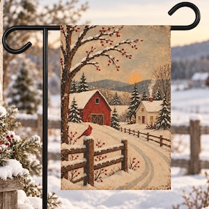 May include: A rectangular garden flag hanging on a black metal stand, featuring a winter landscape illustration with a red barn, a white house, a snow-covered path, a wooden fence, and a bright red cardinal bird perched on a fence post.