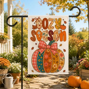 Puede incluir: Una bandera de jardín rectangular con el texto "cozy season" en letras grandes de color naranja y marrón con varios patrones. Debajo del texto hay una ilustración de una calabaza con un lazo a cuadros rojo y blanco, rodeada de hojas de otoño.