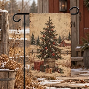 May include: A rectangular garden flag featuring a printed illustration of a decorated Christmas tree in a wooden bucket with wrapped gifts at the base, set in a snowy landscape with a red house and evergreen trees in the background.