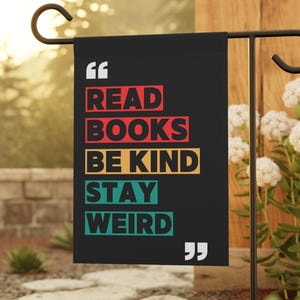 Puede incluir: Una bandera de jardín negra con las palabras "READ BOOKS BE KIND STAY WEIRD" en letras coloridas. La bandera está colgada de un soporte de metal negro en un jardín con flores blancas y piedras.