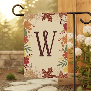 Peut inclure: Un drapeau de jardin rectangulaire avec un fond crème et une lettre W marron foncé au centre. La bordure est ornée d'un motif automnal coloré composé de feuilles rouges, orange, jaunes et vertes, de baies et de petites fleurs.
