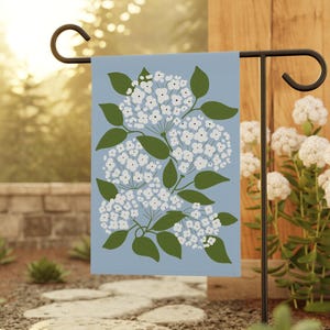 May include: A garden flag featuring a floral design of white hydrangea blooms and green leaves on a light blue background. The flag is displayed on a black metal stand, perfect for outdoor decor.