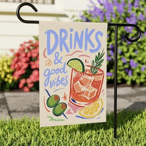 Könnte beinhalten: Eine Gartenflagge mit den Worten "DRINKS & good vibes" in blauer und rosa Schrift. Die Flagge zeigt eine Cocktail-Illustration mit Limette, Oliven und Zitrone. Die Flagge wird auf einem schwarzen Metallständer in einem Garten gezeigt.