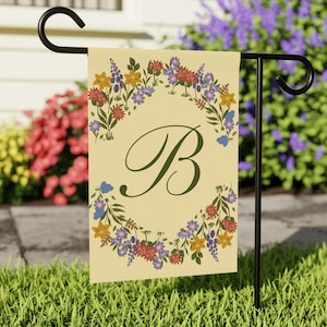 Peut inclure: Un drapeau de jardin rectangulaire au fond jaune clair orné d'une lettre B vert foncé centrale entourée d'une couronne de fleurs sauvages colorées, incluant des pétales rouges, jaunes, violets et bleus avec des feuilles vertes, sur un support en métal noir.