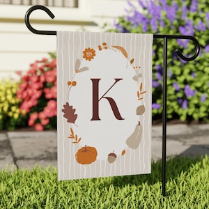 Peut inclure: Un drapeau de jardin rectangulaire avec un fond beige et de fines rayures verticales blanches. Un cadre ovale central contient une grande lettre K marron foncé entourée d'illustrations de citrouilles orange, de glands marron, de feuilles et de fleurs.