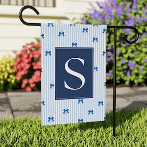 May include: A garden flag with a blue and white striped pattern, adorned with blue bows and a navy blue square containing the letter "S". The flag is displayed on a black metal stand in a garden, with a backdrop of flowers.