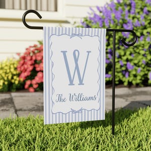 May include: A light blue and white striped garden flag with the letter "W" and the name "The Williams" in a script font. The flag is hanging on a black metal stand in a garden setting.