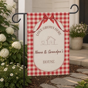 Puede incluir: Bandera de jardín a cuadros rojos y blancos con el texto "LOVE GROWS HERE" y "Nana & Grandpa's HOUSE". La bandera tiene un diseño de cinta roja y un dibujo lineal de una casa con un corazón.
