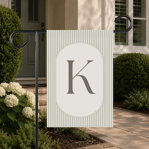 May include: A rectangular garden flag featuring a light green and white vertical striped pattern. In the center, a large, dark gray capital letter K is displayed inside a white oval frame with a thin border, mounted on a black metal stand.