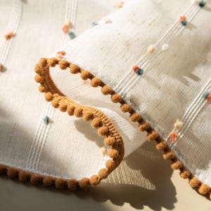 Könnte beinhalten: Nahaufnahme eines gefalteten weißen Stoffes mit strukturiertem Gewebe. Der Stoff ist mit kleinen, farbigen Pompons und einer braunen Pomponborte verziert. Vertikale Streifen verleihen dem Textil ein subtiles Muster und schaffen eine weiche, einladende Ästhetik.