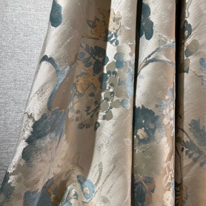 May include: A close-up of a beige fabric with a blue and gold floral pattern. The fabric has a textured surface and a subtle sheen.