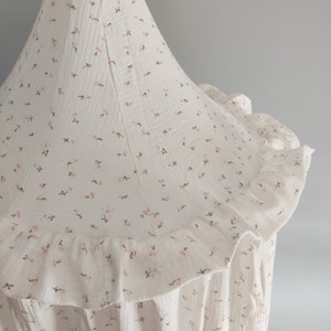 May include: A white canopy with a floral pattern of small pink and green flowers. The canopy has a ruffled edge and is made of a soft, textured fabric. The design is delicate and suitable for a nursery or children's room.