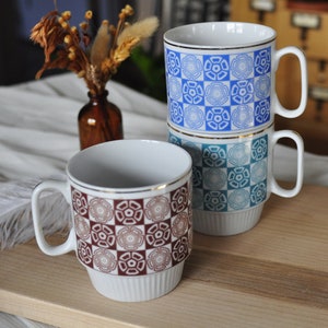 May include: Three ceramic mugs with geometric patterns. The mugs are stacked on a wooden surface. The top mug is blue and white, the middle mug is blue and green, and the bottom mug is brown and white.