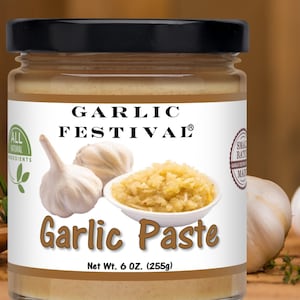 May include: A jar of Garlic Festival garlic paste with a black lid. The label features garlic bulbs and a bowl of minced garlic. The jar contains 6 oz (255g) of garlic paste. Fresh garlic bulbs are arranged around the jar.