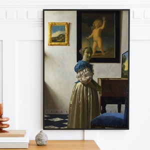 May include: A framed painting of a woman in a blue and white dress standing in front of a window. The woman is looking to the right and holding a book. The painting is set against a white wall with a smaller framed painting above it.