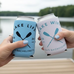 May include: Two can coolers, one light blue and one white, are held up in front of a lake. The light blue cooler says "Lake Life" with crossed oars and "Mia's Bachelorette". The white cooler says "Lake Wife" with crossed oars and "Mia's Bachelorette".