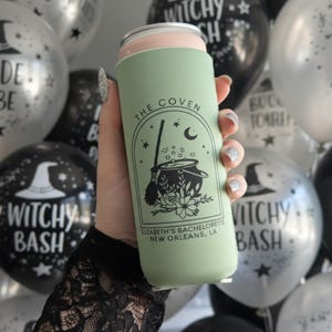May include: A light green can cooler with a black witch-themed design, including a cauldron, broom, and text that reads "The Coven" and "Elizabeth's Bachelorette New Orleans, LA." The background features black and silver balloons with witch hat and "Bride to Be" text.