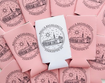 May include: Pink can coolers with a black and white design featuring a city skyline and the text "MOLLI'S BACHELORETTE CHARLESTON, SC".