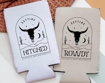 May include: Two white and beige can coolers with black text and graphics. The white cooler says "Getting Hitched Olivia's Bachelorette" and the beige cooler says "Getting Rowdy Olivia's Bachelorette". Both coolers feature a black skull with horns and a crescent moon in a desert landscape.