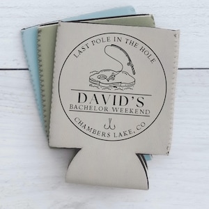 May include: A white can cooler with a black circle design. The circle says "Last Pole in the Hole" with a fishing rod and fish inside. Below the circle it says "David's Bachelor Weekend Chambers Lake, CO".