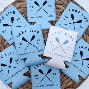 May include: A collection of light blue and white drink koozies with the text "LAKE LIFE" and "LAKE WIFE". Each koozie features crossed oars, the number "20" and "26", and "MIA'S BACHELORETTE". The koozies are arranged on a woven surface.