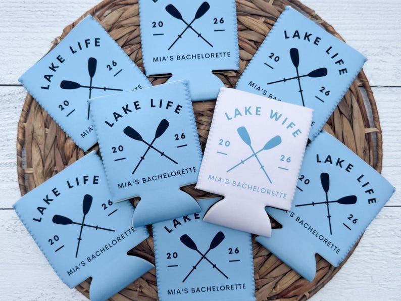May include: A collection of light blue and white drink koozies with the text "LAKE LIFE" and "LAKE WIFE". Each koozie features crossed oars, the number "20" and "26", and "MIA'S BACHELORETTE". The koozies are arranged on a woven surface.