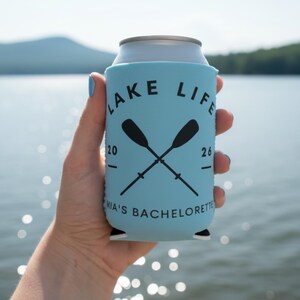 May include: A light blue can cooler with the words "LAKE LIFE" in black, along with crossed oars. The text "MIA'S BACHELORETTE" is printed at the bottom. The cooler is held in front of a lake.