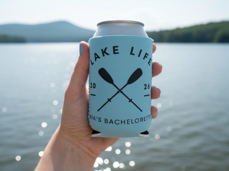 May include: A light blue can cooler with the words "LAKE LIFE" in black, along with crossed oars. The text "MIA'S BACHELORETTE" is printed at the bottom. The cooler is held in front of a lake.