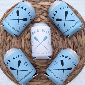 May include: A collection of blue and white can coolers with the text "LAKE LIFE" and "LAKE WIFE". Each cooler features crossed oars and the text "MIA'S BACHELORETTE". The coolers are arranged on a woven wicker placemat.