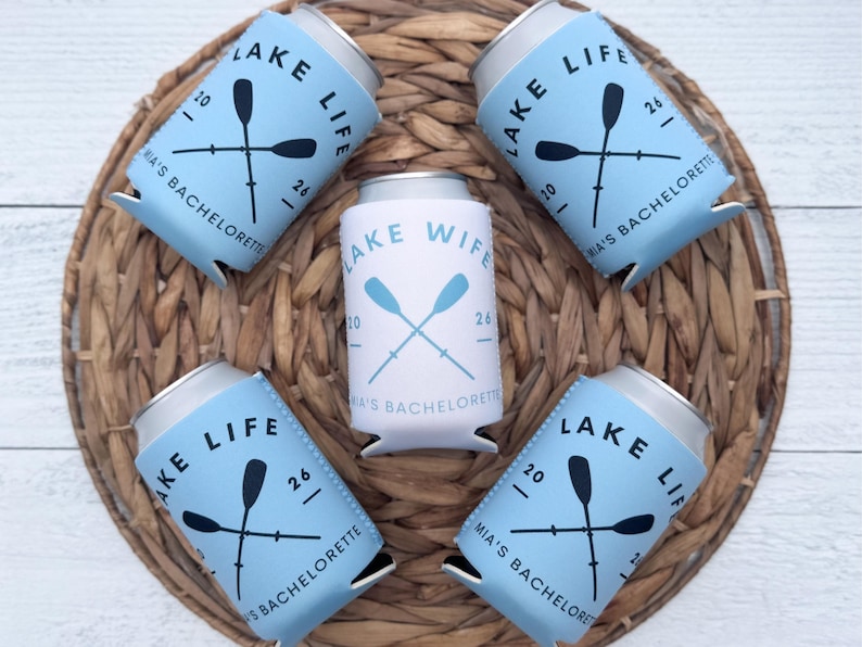 May include: A collection of blue and white can coolers with the text "LAKE LIFE" and "LAKE WIFE". Each cooler features crossed oars and the text "MIA'S BACHELORETTE". The coolers are arranged on a woven wicker placemat.
