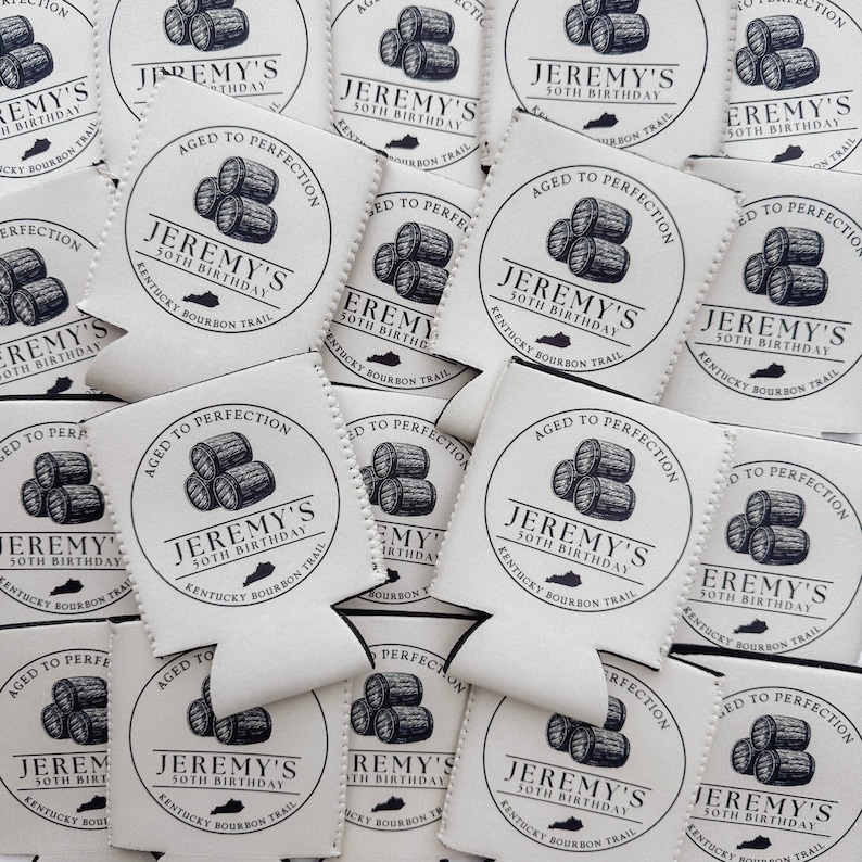 Pu&ograve; includere: Una pila di refrigeratori per lattine bianchi con un designo in bianco e nero. Il design presenta un cerchio con il testo "Jeremy's 50th Birthday" e una grafica di tre barili. Il testo "Kentucky Bourbon Trail" &egrave; incluso anche nel design.