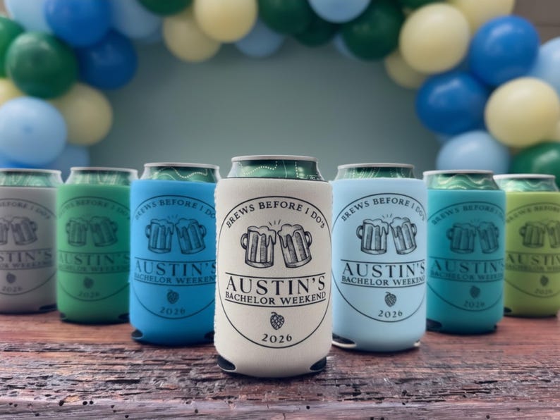 May include: A collection of beer can coolers in various colors, including gray, green, blue, and white. Each cooler features the text "BREWS BEFORE I DO" and "AUSTIN'S BACHELOR WEEKEND 2026" with a beer mug graphic. The background includes balloons.