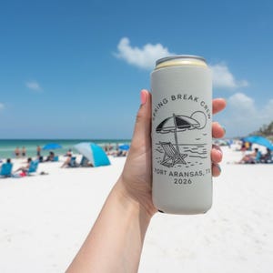 Puede incluir: Un enfriador de latas gris claro con el texto "Spring Break Crew Port Aransas, TX 2026" y un gr&aacute;fico de escena de playa. El enfriador se sostiene contra un fondo de playa soleado con gente y sombrillas.