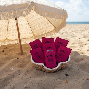 Puede incluir: Una escena de playa con una sombrilla crema con flecos que proyecta sombra sobre una cesta llena de enfriadores de latas rosas. Los enfriadores y una lata est&aacute;n impresos con un logotipo con tem&aacute;tica de playa. La escena est&aacute; ambientada en una playa arenosa con el oc&eacute;ano al fondo.