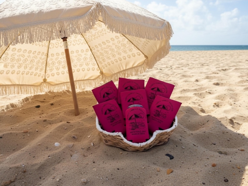 Puede incluir: Una escena de playa con una sombrilla crema con flecos que proyecta sombra sobre una cesta llena de enfriadores de latas rosas. Los enfriadores y una lata est&aacute;n impresos con un logotipo con tem&aacute;tica de playa. La escena est&aacute; ambientada en una playa arenosa con el oc&eacute;ano al fondo.