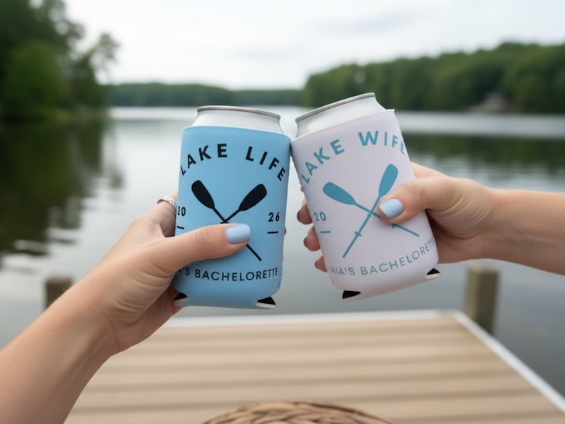 May include: Two can coolers, one light blue and one white, are held up in front of a lake. The light blue cooler says "Lake Life" with crossed oars and "Mia's Bachelorette". The white cooler says "Lake Wife" with crossed oars and "Mia's Bachelorette".