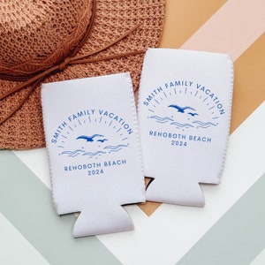 K&ouml;nnte beinhalten: Zwei wei&szlig;e Schaumstoff-Getr&auml;nkedosenk&uuml;hler mit blauem Text und Grafiken. Der Text lautet "Smith Family Vacation" und "Rehoboth Beach 2024". Die Grafik zeigt eine Sonne mit Strahlen und zwei M&ouml;wen, die &uuml;ber Wellen fliegen.