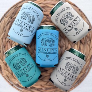 May include: Five beer can coolers in shades of blue, teal, and gray, arranged on a woven mat. Each cooler features the text "Brews Before I Do's" and "Austin's Bachelor Weekend 2026" with a beer mug graphic. The cans are visible at the top.
