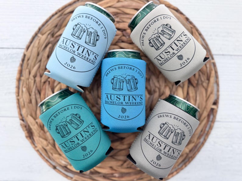 May include: Five beer can coolers in shades of blue, teal, and gray, arranged on a woven mat. Each cooler features the text "Brews Before I Do's" and "Austin's Bachelor Weekend 2026" with a beer mug graphic. The cans are visible at the top.