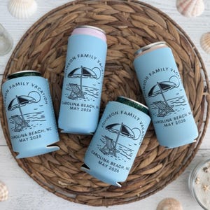 Puede incluir: Cuatro enfriadores de latas azul claro con un dise&ntilde;o de playa. Cada enfriador presenta una ilustraci&oacute;n en blanco y negro de una sombrilla, una silla y el texto "JOHNSON FAMILY VACATION CAROLINA BEACH, NC MAY 2026". Los enfriadores est&aacute;n colocados sobre una estera tejida con conchas marinas.