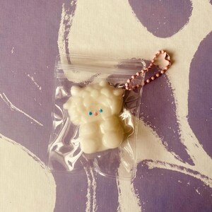 May include: A white, fluffy, cartoon-like figure with blue eyes and a pink chain keychain is in a clear plastic bag. The background has a purple and white abstract pattern.